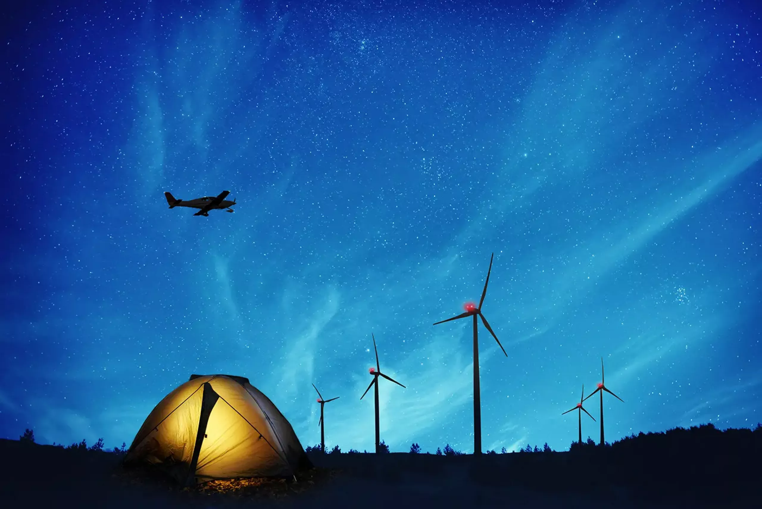 Ein beleuchtetes Zelt unter einem sternenklaren Nachthimmel, mit Windenergieanlagen, die mit BNK ausgestattet sind und einem darüber fliegenden Flugzeug im Hintergrund.
