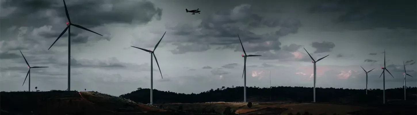 Ein mit BNK ausgestatteter Windpark mit mehreren Windturbinen und einem darüber fliegenden Flugzeug vor einem Hintergrund von dunklem, bewölktem Wetter.