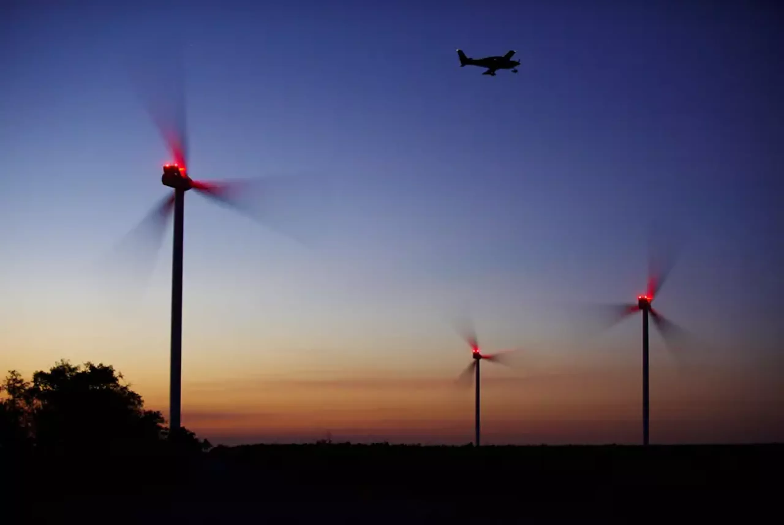 Drei Windenergieanlagen mit roten Lichtern sind durch bedarfsgesteuerter Nachtkennzeichnung bei Dämmerung beleuchtet, während ein Flugzeug im dunkler werdenden Himmel darüber fliegt.