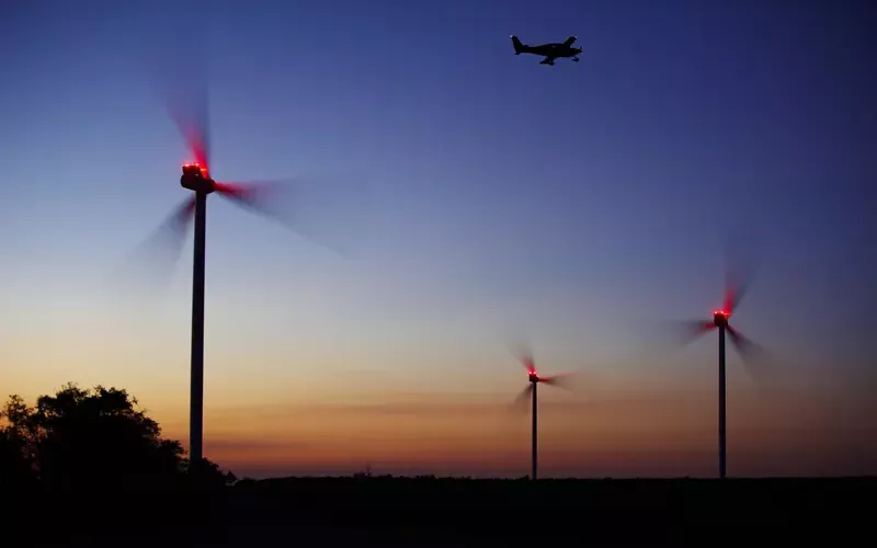 Drei Windenergieanlagen mit roten Lichtern sind durch bedarfsgesteuerter Nachtkennzeichnung bei Dämmerung beleuchtet, während ein Flugzeug im dunkler werdenden Himmel darüber fliegt.