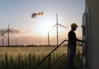 Ein Techniker steht auf einer Plattform und inspiziert die bedarfsgesteuerte Nachtkennzeichnung von Windenergieanlagen bei Sonnenuntergang, während ein Hubschrauber im Hintergrund fliegt. Im Hintergrund sind weitere Windenergieanlagenzu sehen.