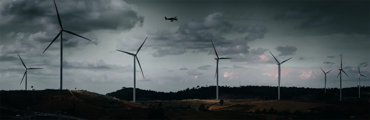 A wind farm equipped with ADLS with several wind turbines and an aeroplane flying above against a background of dark, cloudy weather.